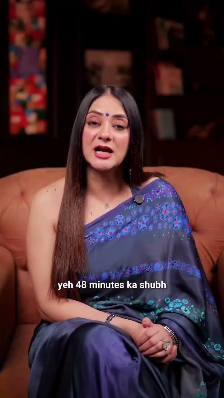 A woman in a blue and purple saree seated indoors, with a subtitle across the frame